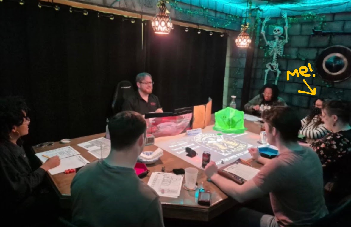 Lissy and the adventuring party sitting at a massive table facing the gamemaster in a dark, fantasy theme room